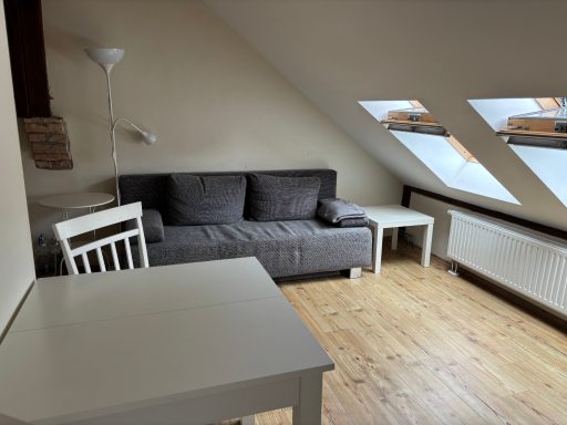 Ein sonniger Dachzimmer mit einer grauen Couch, zwei Tischen und Dachfenstern.