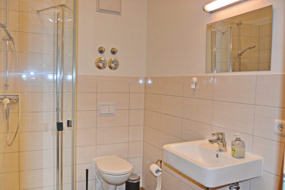 Helles Badezimmer mit Dusche, Waschbecken und Toilette, helle Fliesen und Wandspiegel.