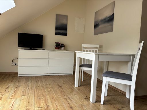 Helles Zimmer mit Holzfußboden, TV, Kommode und Tisch mit Stühlen.