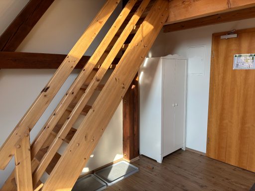 Holztreppe zu einem oberen Stockwerk, Kühlschrank und Tür im Raum sichtbar.