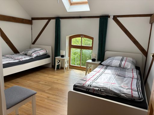 Schlafzimmer mit zwei Betten, Fenster mit Blick ins Grüne, Holzboden und einfache Möbel.